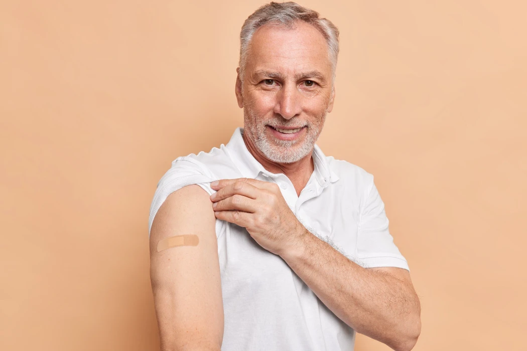Old bearded man got vaccinated against coronavirus shows arms with adhesive plaster cares about healthy during pandemic poses against brown wall